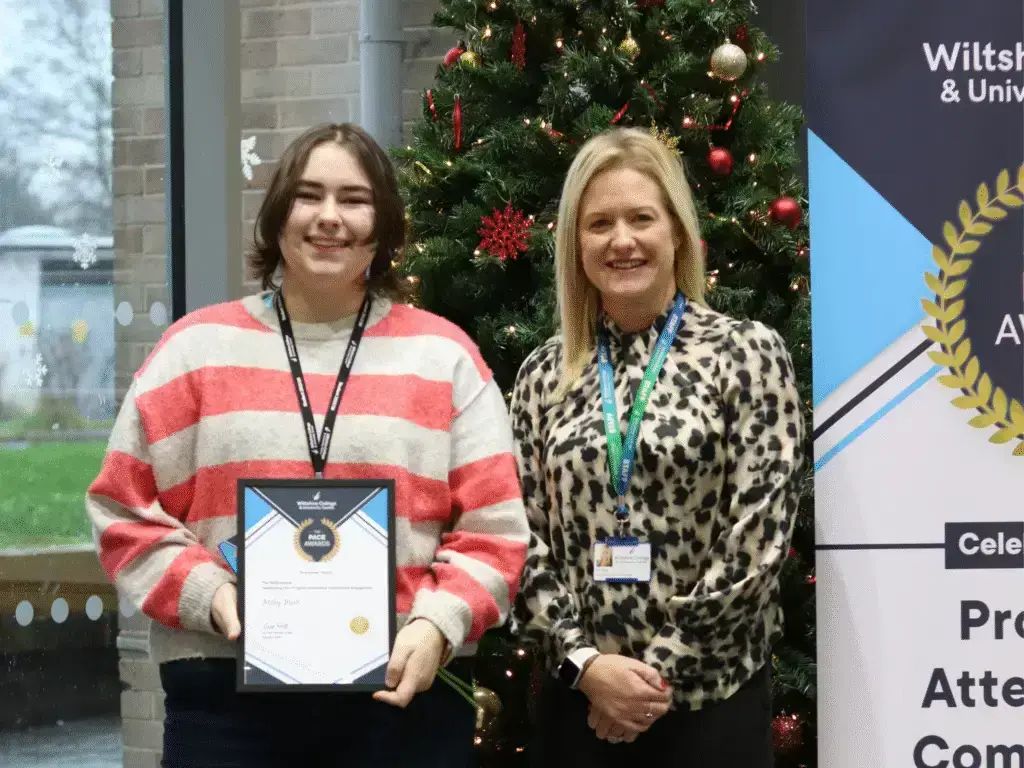 A student holding a PACE Award certificate stands beside a member of staff in front of a Christmas tree and a Wiltshire College & University Centre PACE Awards banner.