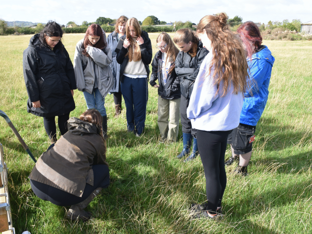 Students gather around a soil sampling demonstration in a grassy field.