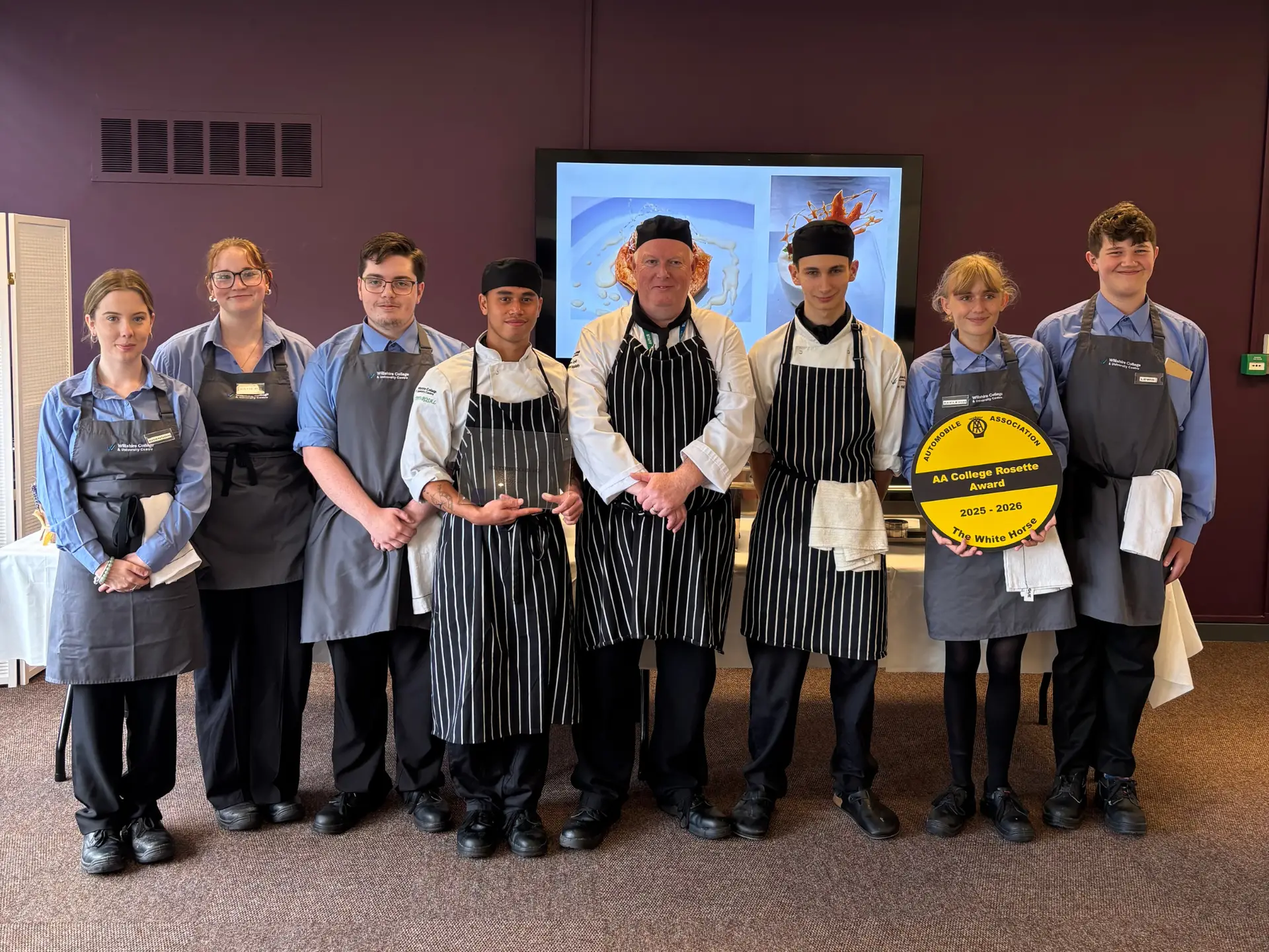 Hospitality students and staff stand together in uniform, with one student holding an AA College Rosette Award plaque.