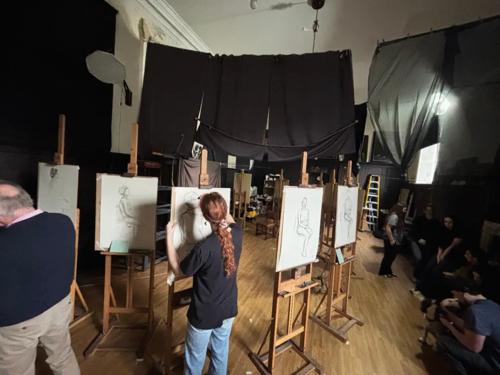 Students positioned at individual easels during a structured life drawing class at Sarum Studio, with natural light and draped studio backdrops visible.
