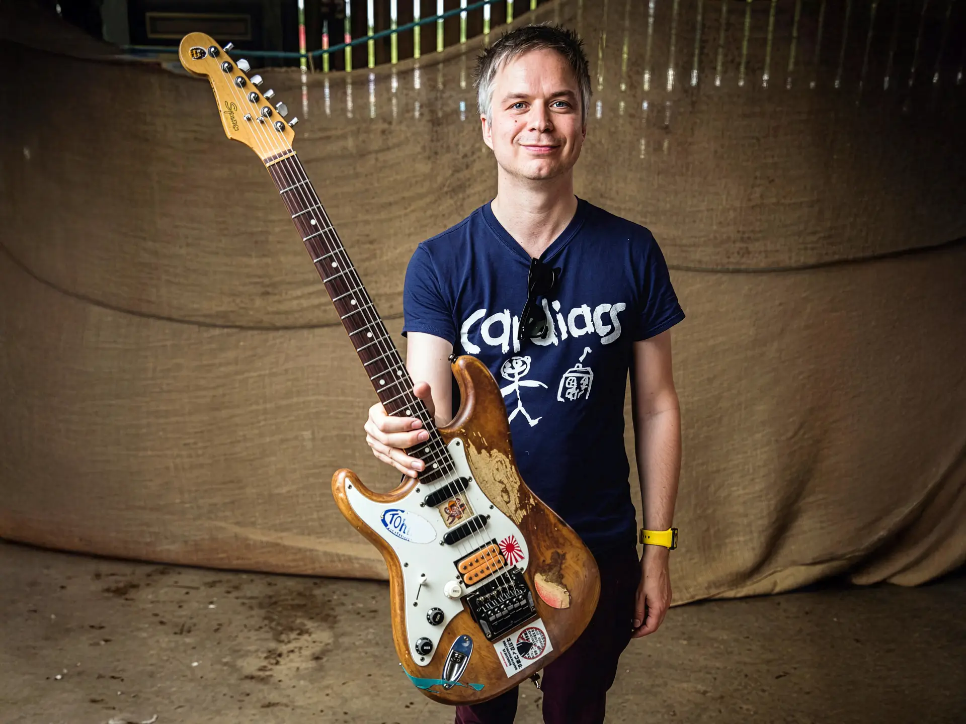 A musician holding an electric guitar, standing indoors and smiling at the camera.