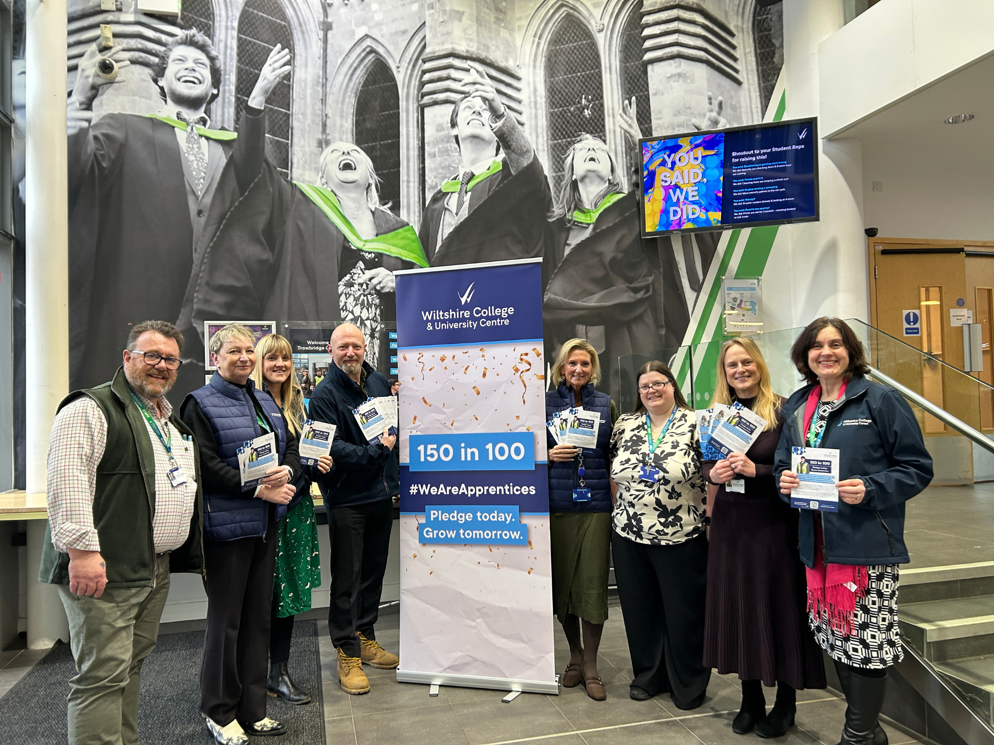 Staff holding 150 in 100 campaign leaflets beside a Wiltshire College & University Centre banner in the campus atrium.