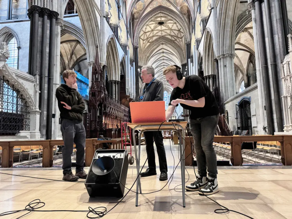 Music students and a lecturer set up recording equipment and a laptop inside Salisbury Cathedral, capturing the building’s acoustics using a speaker and microphones.