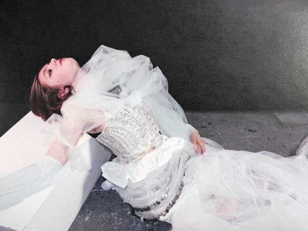 Model sat with her back arched and looking up in a white lacey dress 