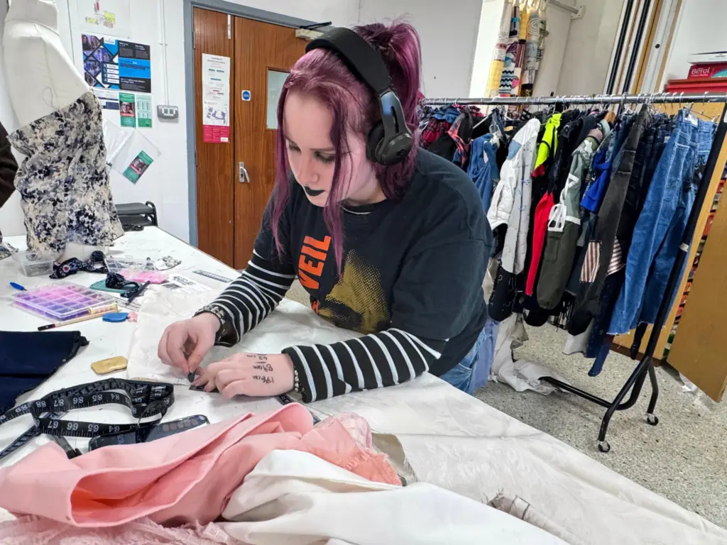 Fashion Student at WCUC working on clothing on a table with a rack of clothes behind her.