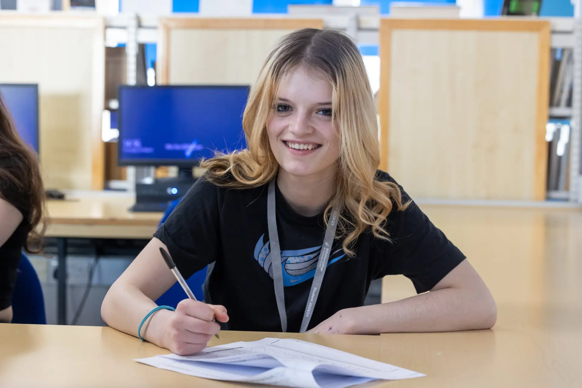 Student sat at a desk holding a pen with paper in front of her. She looks at the camera and smiles.