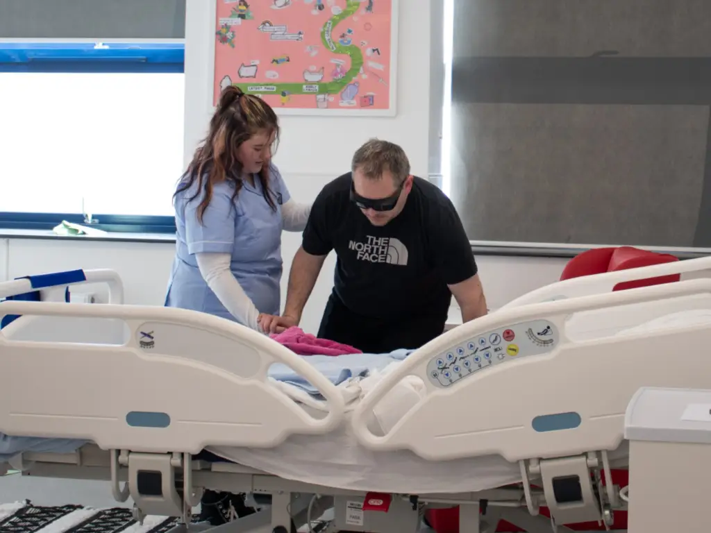 A health and social care student helping a gentleman on to a hospital bed