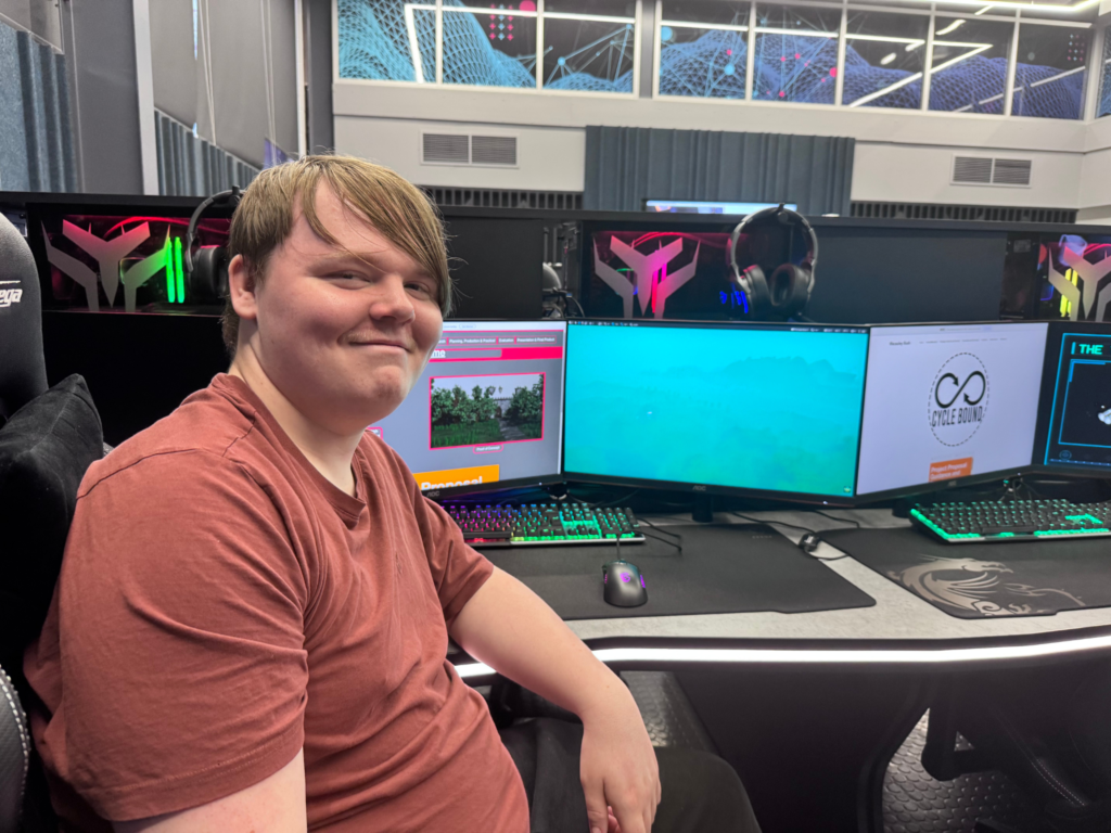 Student seated beside a games development project display featuring digital artwork and computer screens.