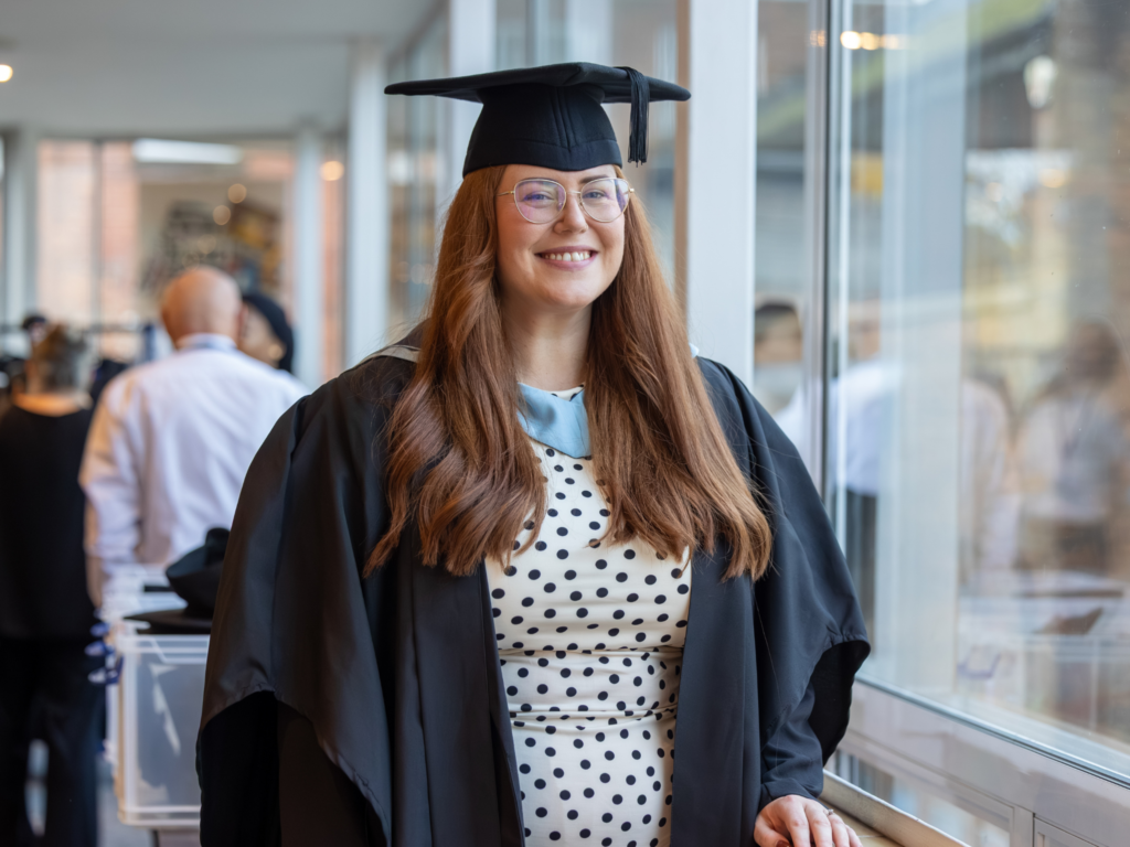A graduand stood by a window in gown and cap 